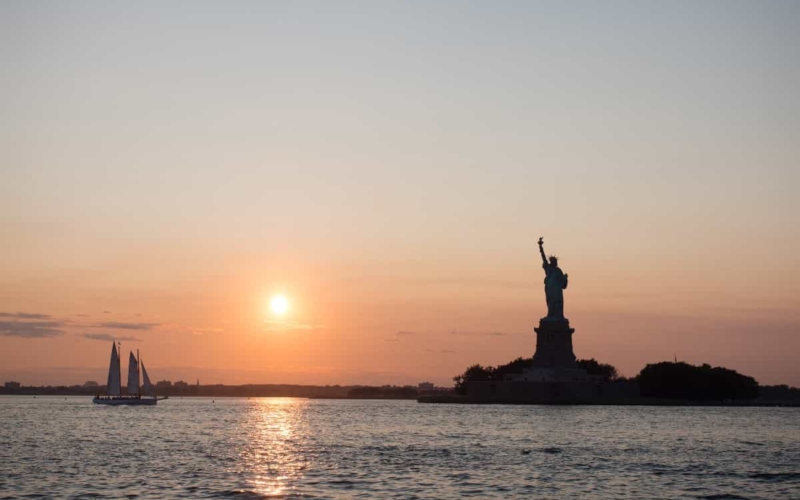 sailboat next to statue of liberty at sunset