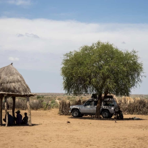 off road vehicle parked under tree next to tribe members seeking shade under hut