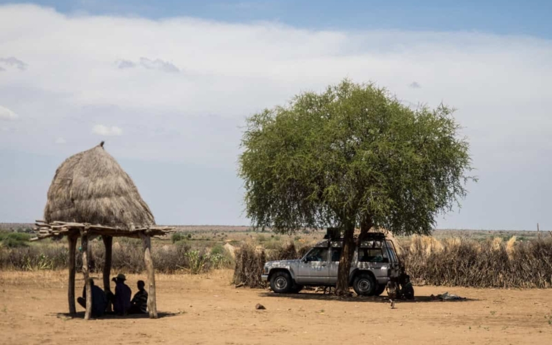 off road vehicle parked under tree next to tribe members seeking shade under hut