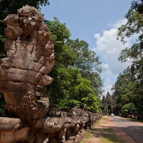 Ancient stone serpent sculptures line a path surrounded by trees, leading toward the entrance of a temple in the distance under a partly cloudy sky.