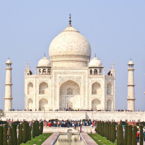 The image shows the front view of the Taj Mahal, with crowds of visitors, reflecting pool, and four minarets under a clear sky.