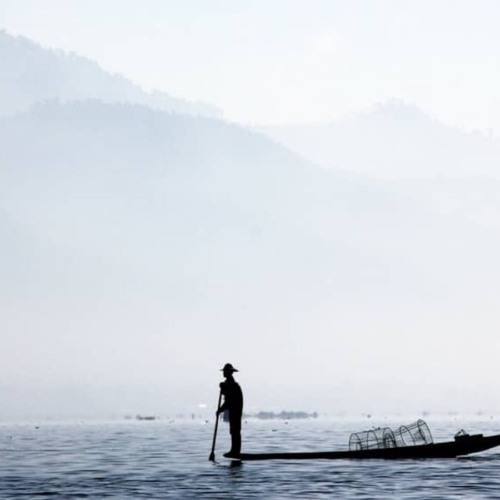 A person stands on a narrow boat with a paddle in hand on a calm body of water, with mist-covered mountains in the background.