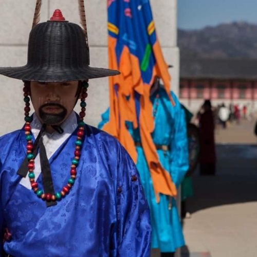 A guard in traditional blue attire and black hat stands at a palace entrance, with more guards and colorful flags in the background.