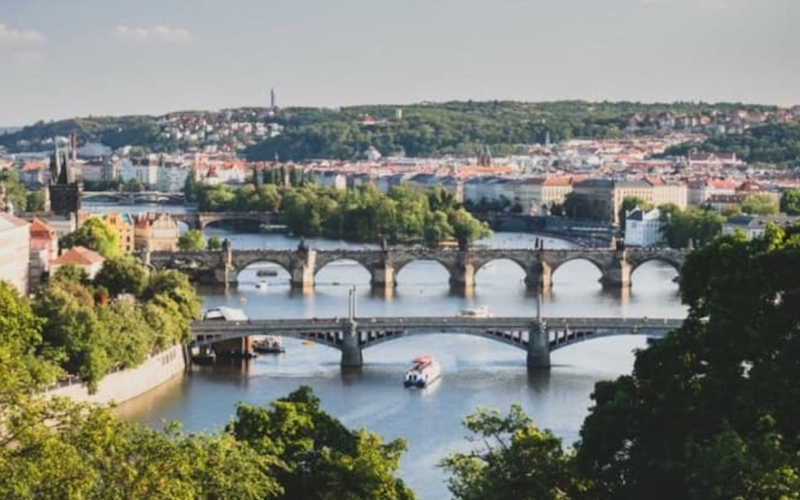 A scenic view of multiple historic bridges crossing a river in a city, with buildings and green hills in the background under a partly cloudy sky.