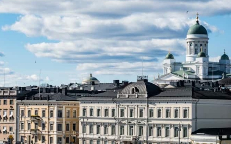 Cityscape view featuring neoclassical buildings and the white Helsinki Cathedral with green domes under a partly cloudy sky.