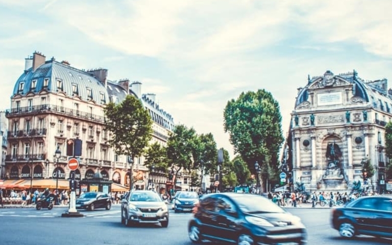 Cars drive through a busy intersection in a European city, with historic buildings, shops, trees, and a large stone fountain in the background.