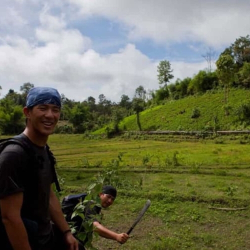 Two people outdoors in a green, grassy field; one is smiling at the camera with a backpack, while the other holds a machete and crouches near the ground.