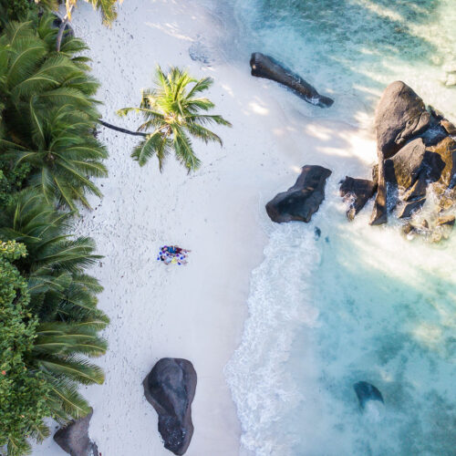 Aerial view of a tropical beach with white sand, palm trees, large rocks, and clear turquoise water; a small group of people is lying on the sand near the shoreline.